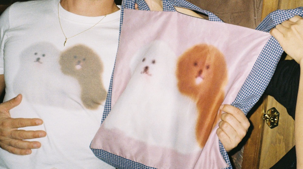 a friend holding up a pink tote bag printed with two fluffy dogs, next to a white t-shirt with the same dogs