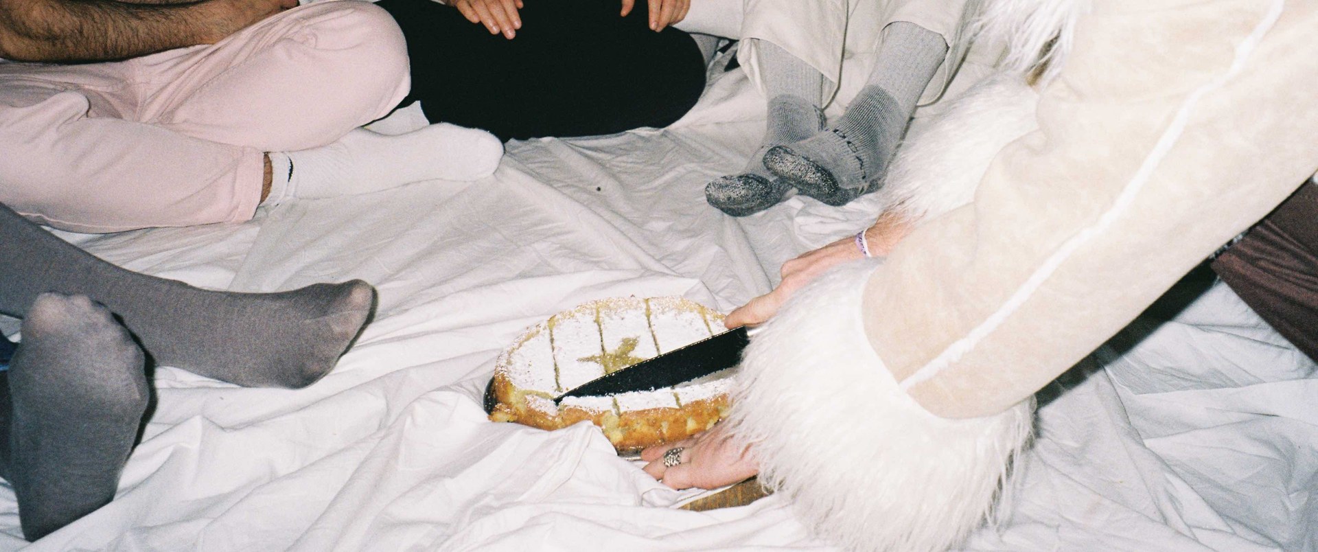 friends sitting on a white sheet on the floor, one of them cutting a cake dusted with sugar